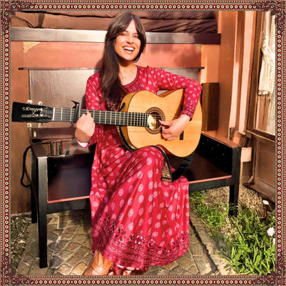 Woman wearing the Rani Rouge Jaipur Kurta Dress by FAVIE while sitting with a classical guitar, showcasing the flowing red-pink bohemian maxi dress.